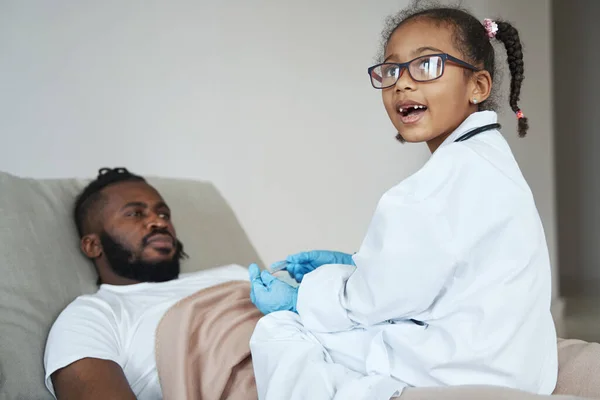 Ragazza afroamericana in abito medico si siede su suo padre con un termometro — Foto Stock