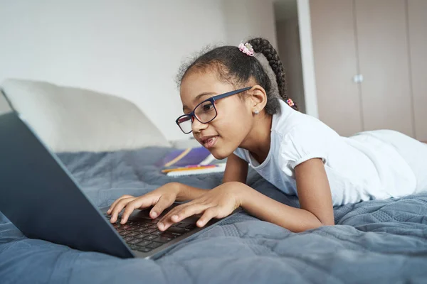 Little African American girl on bed looking at laptop screen — Stock Photo, Image