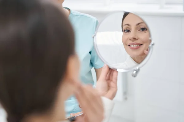Cheerful woman looking in the mirror in cosmetology clinic — Stock Photo, Image