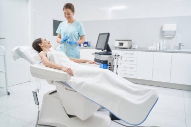 Neşeli güzellik uzmanı kozmetoloji kliniğinde bir kadınla konuşuyor.