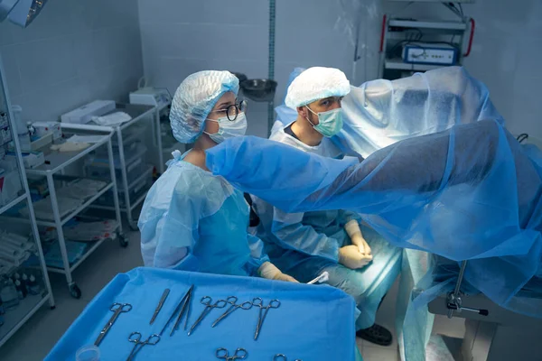 Surgeons sitting near lifted legs of patient on operating table — Stock Photo, Image