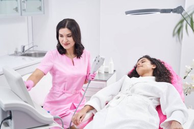 Tatmin olmuş bayan tıbbi muayenehanede yatarken doktor lazer tüy temizleyiciyi hazırlıyor.