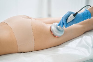 Kozmetik uzmanı selülit çıkarma sırasında uyluk derisini ısıtmak için el dalgası kaldırma makinesi kullanıyor.