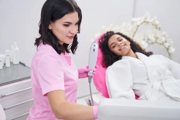 Attentive Cosmetologist Studying Settings Medical Device While Young Woman Lying — Stock Photo, Image