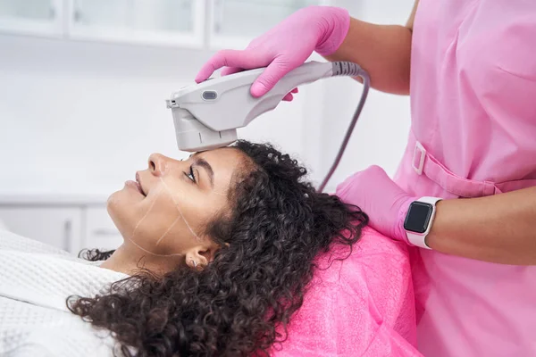 リラックスした女性は 医師が機械で化粧品の手順を実行しながら 彼女の顔にマーキングに横たわっています — ストック写真