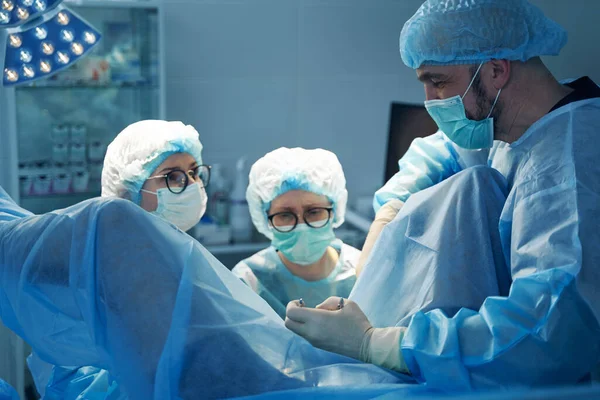 Cirurgião Masculino Segurando Grampos Enquanto Duas Enfermeiras Realizando Operação Uma — Fotografia de Stock