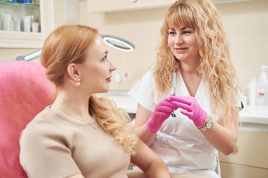 Mutluluktan gülümseyen doktorun kozmetik enjeksiyonlu belden yukarısı klinikteki kadın müşteriyle konuşuyor.