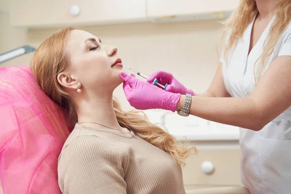 Kozmetik salonundaki bayana hyaluronik dudak enjeksiyonu yapan tanınmamış bir doktorun belden yukarı portresi.