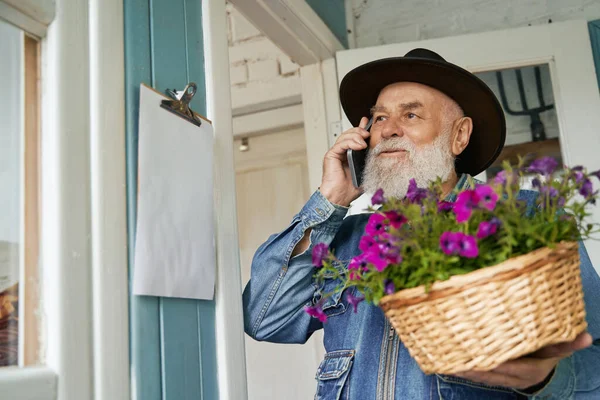 카우보이 모자를 핸드폰으로 말하고 보라색 꽃이든 바구니를 다니는 노인의 — 스톡 사진