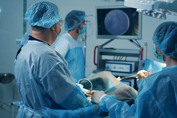 Doctors in medical masks and surgical gowns looking at the screen while using hysteroscope in the operating room