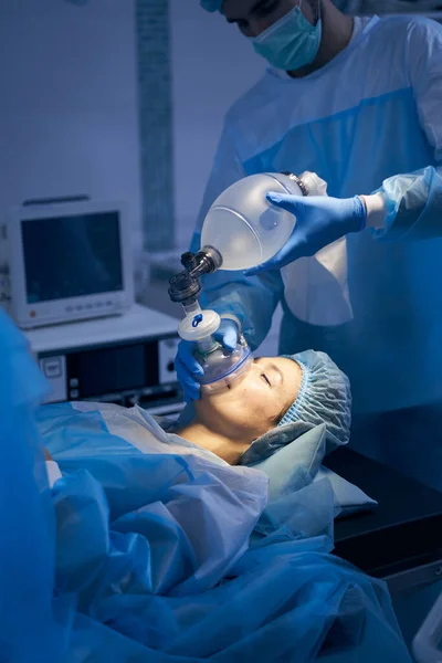 Anesthesiologist wearing surgical gown and medical mask standing behind the patient and maintaining anesthesia