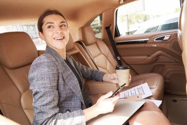 Gülümseyen genç bir kadın otomobilde iş gezisi yaparken elinde bir fincan kahve ve akıllı telefon tutuyor.