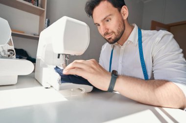 Düşük açılı, rahat bir genç adam yan masada oturuyor ve elbise dikmek için dikiş makinesi kullanıyor.