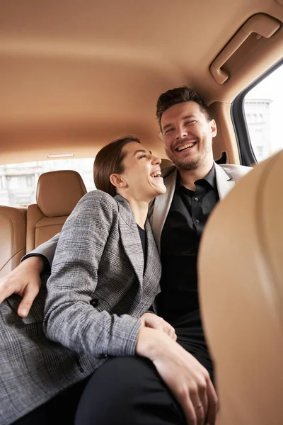 Cheerful businessman hugging woman and smiling while sitting on back seat in car