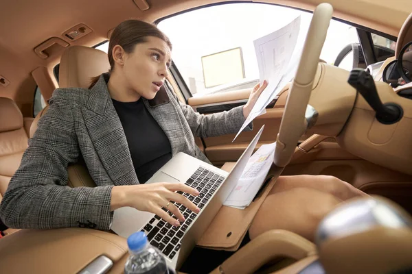 Businesswoman having phone conversation and studying documents while working on laptop in automobile