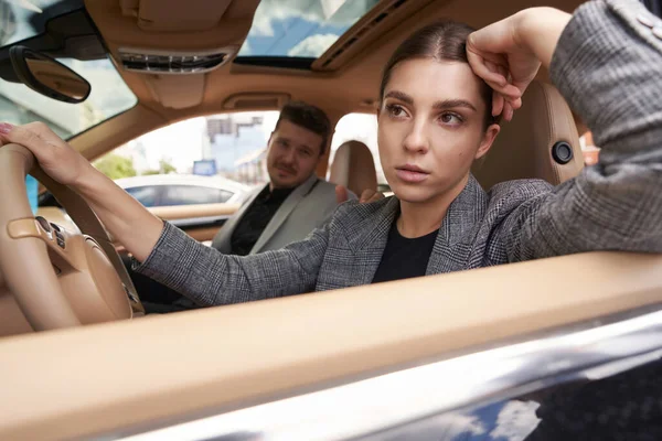 Upset lady sitting on driver seat with her hand on steering wheel and looking out car window while having argument with husband