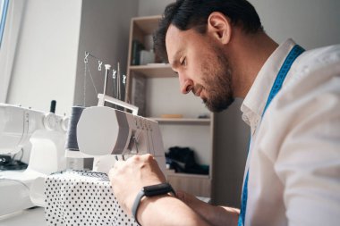 Düşük açılı genç adam masada oturuyor ve polka noktalı kumaşla çalışırken dikiş makinesi kullanıyor.