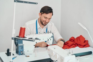 Mutlu genç adam modern ekipmanlarla masada oturuyor ve atölyede kırmızı elbiseler yapıyor.