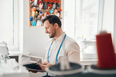 Elinde bant olan gülümseyen erkek terzi atölyede duruyor ve elinde kumaş örnekleriyle dergi tutuyor.