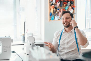 Neşeli adam iplik makaralarının yanında oturuyor ve dikiş makinesinde cep telefonuyla konuşuyor.