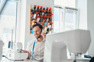 Sandalyede cep telefonuyla konuşurken dikiş makinesi kullanan neşeli terzinin düşük açısı