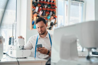 Düşük açılı terzi sandalyede oturmuş dikiş makinesi ve cep telefonuyla mesajlaşıyor.