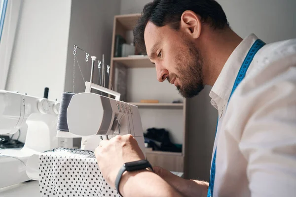 Düşük açılı genç adam masada oturuyor ve polka noktalı kumaşla çalışırken dikiş makinesi kullanıyor.