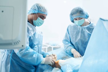 Tıbbi maskeli iki cerrah bacağını dikkatlice ameliyat ediyor. Ellerinde neşter ve kıskaçlar