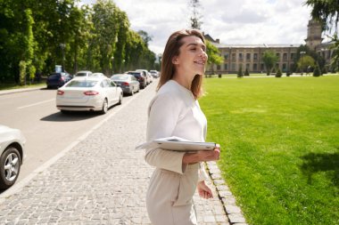 Şehirde dışarıda vakit geçirirken elinde dizüstü bilgisayar ve belgelerle gülümseyen kadın.