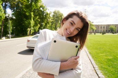 Kameraya bakan, elinde dizüstü bilgisayarla gülümseyen ve dışarıda doküman tutan neşeli bir kadın.