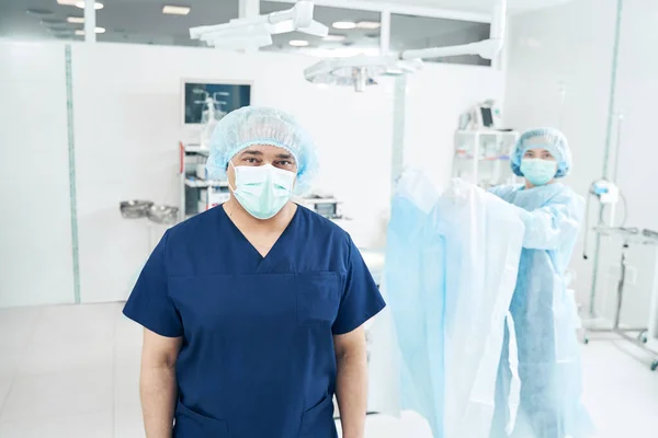 Professional male surgeon standing in the operation room with a nurse holding protection suit