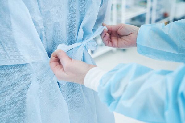 Close up of medical assistant tying a ribbon of the surgical gown of his colleague