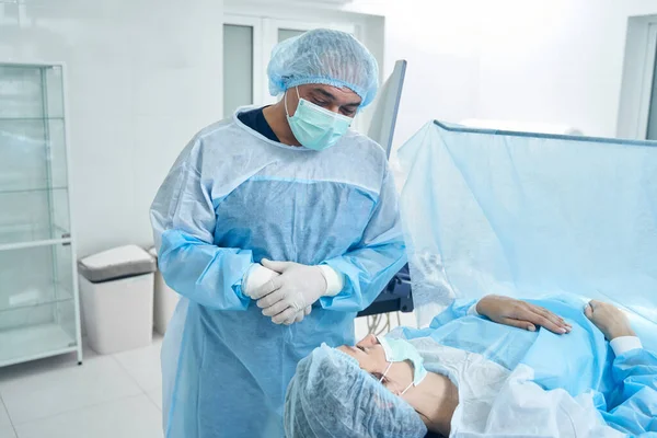 Male doctor in surgical gown looking at the patient lying on the operation table behind the surgical screen