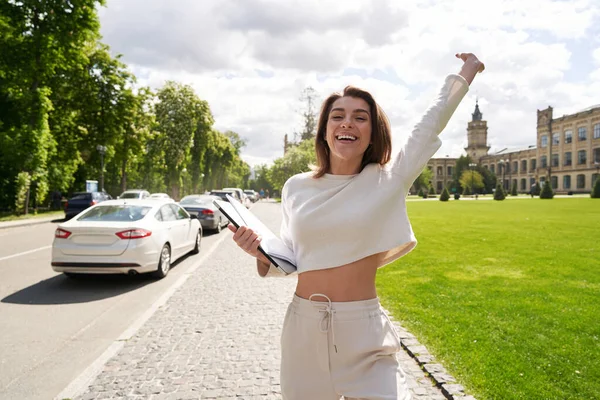 Joyful excited lady holding notebook and papers while raising hand and smiling with joy outdoors