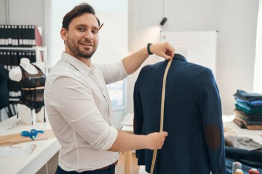 Neşeli moda tasarımcısı, elleri ceketin uzunluğunu ölçmek için bant koyarken mankenin yanındaki dikiş atölyesinde duruyor.