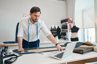 Ciddi bir moda tasarımcısı dizüstü bilgisayara bakarken yanında dikiş dikmek için hazır bir kumaş parçası duruyor.