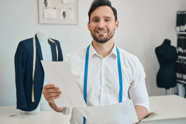 Cheerful fashion designer is standing near the desktop while his hands are holding documents