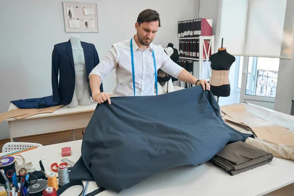 stock image Focused fashion designer stands near the desktop in sewing workshop while unfolding a roll of fabric to its full width