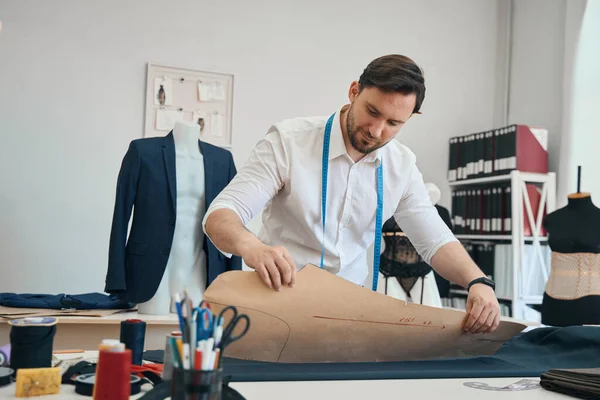 Calm fashion designer is applying blank of future clothes to fabric while behind him stands a mannequin with a jacket