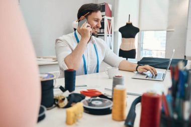 Telefon görüşmeleri sırasında dizüstü bilgisayara bakan tasarımcı önündeki masaya dikiş ekipmanları seriliyor.