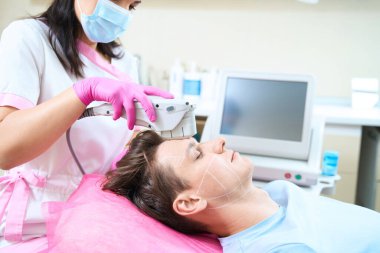 Erkek müşteri güzellik kliniğinde kanepede yatarken kadın profesyonel kırışıklıkları önlemek için modern teknolojiyi kullanıyor.