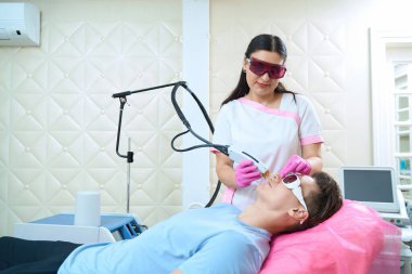 Gülümseyen kozmetikçi, erkek müşteriyle kanepede duruyor ve yüzüne lazer sürüyor.