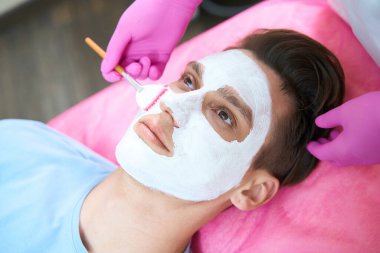 Güzellik uzmanları maske takmak için fırça kullanırken genç bir erkeğin kanepede yatışının üst görüntüsü.