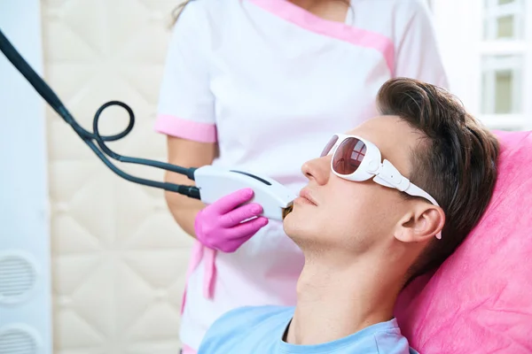 Calm male in protective glasses in lying in couch while cosmetologist is removing hair with laser