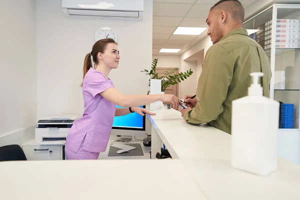 Woman medical worker standing behind reception desk and pointing at clipboard while man filling out patient documents