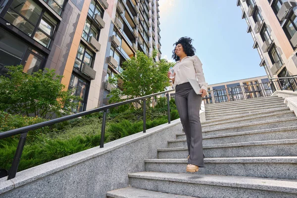 Smiling multiracial woman walking up stairs and holding laptop and water
