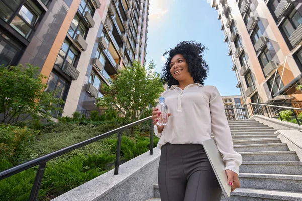 Happy woman goes down the stairs on a sunny day and holds a laptop and water in her hands