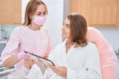 Doktor mücevher kataloğunu gösterirken, mutlu bayan da elinde aksesuarlarla tıbbi kanepede oturuyor.