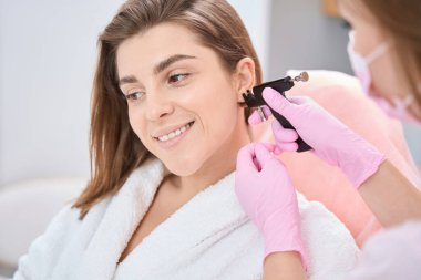 Smiling lady is sitting in bathrobe near beautician while aesthetic medicine doctor put piercing device to her ear