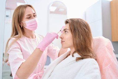 Focused lady looking straight in beauty parlor while doctor doing cosmetic procedure